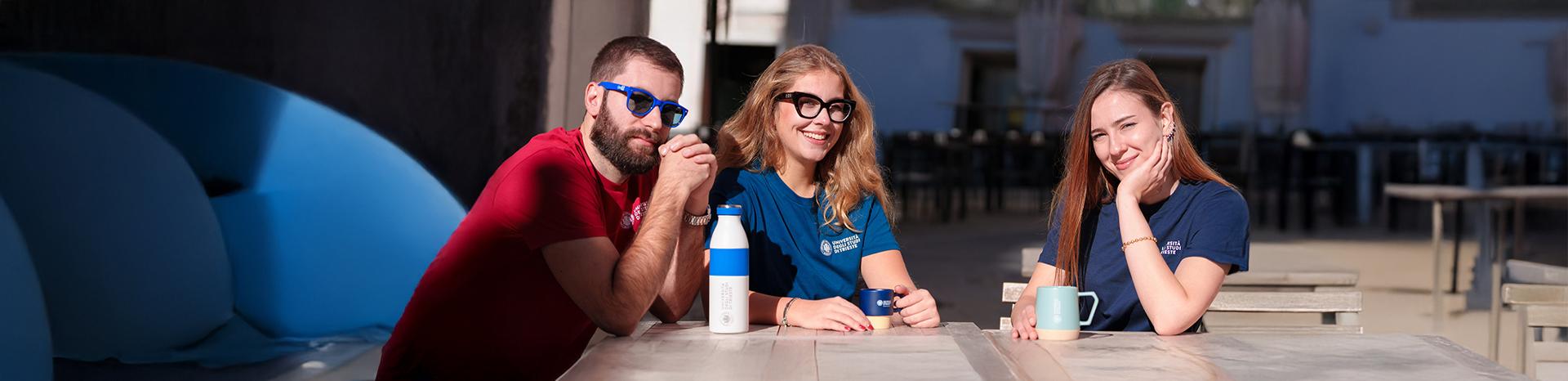 tre studenti con vestiario e gadget UniTS seduti a un tavolo